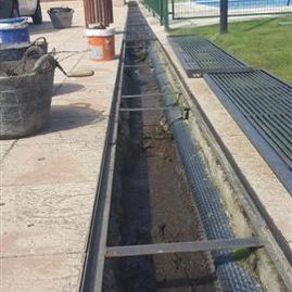 Zanja abierta junto a un paseo con rejillas metálicas, conos y cubos de obra en un muelle o en la cubierta de una piscina