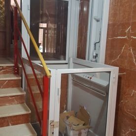 Escalera interior con un pequeño ascensor blanco o montaplatos junto a barandillas rojas y paredes de mármol