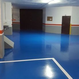 Pasillo interior vacío, con el suelo azul brillante, paredes grises y una puerta oscura al fondo.