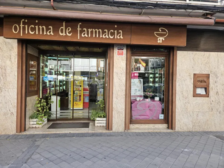 Farmacia a pie de calle con letrero marrón, puertas de cristal y escaparate en la acera.