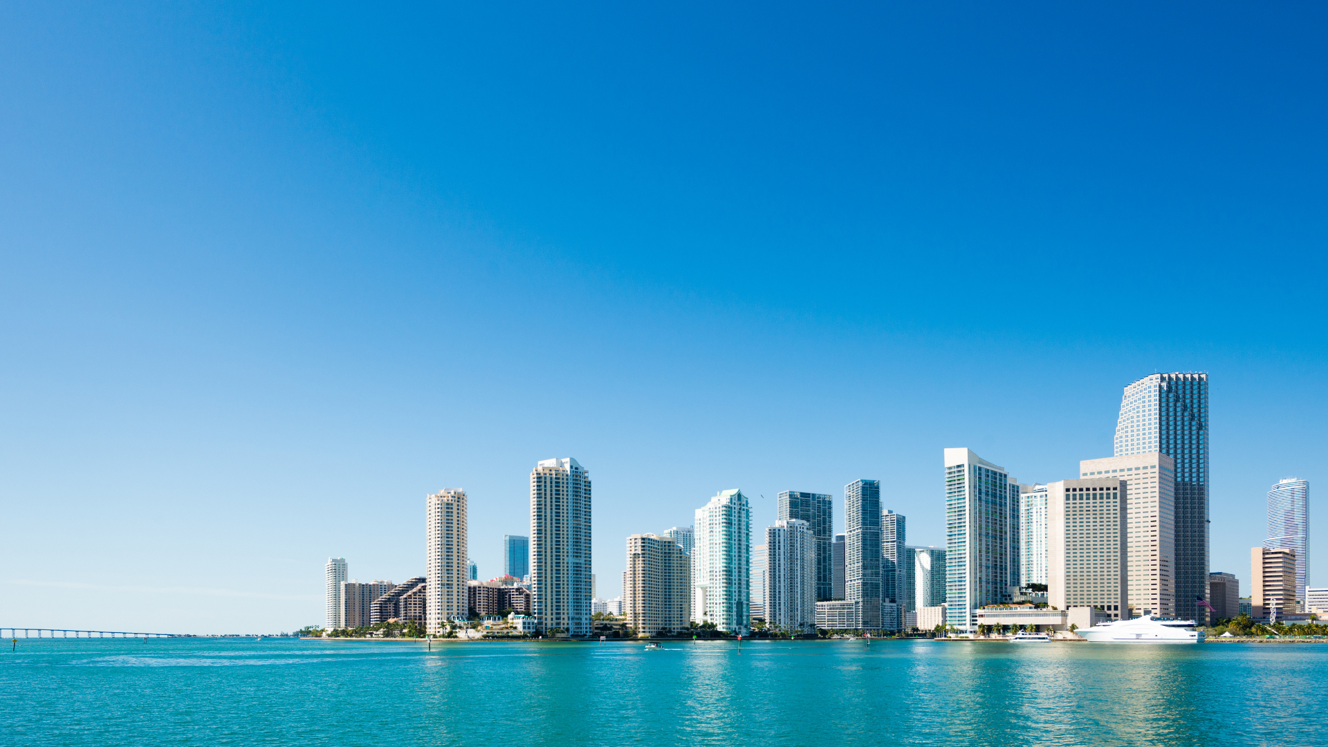 Miami skyline against a bright blue sky, viewed over turquoise water.