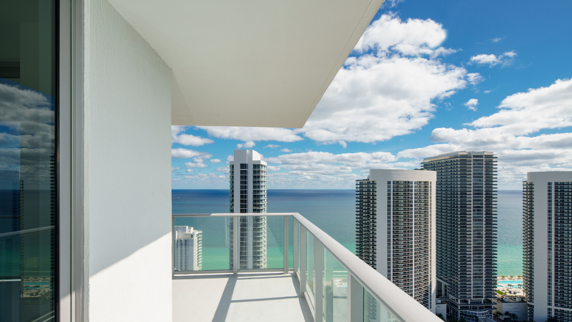 Ocean view from a high-rise balcony with glass railings, bright blue sky, clouds, and tall buildings.