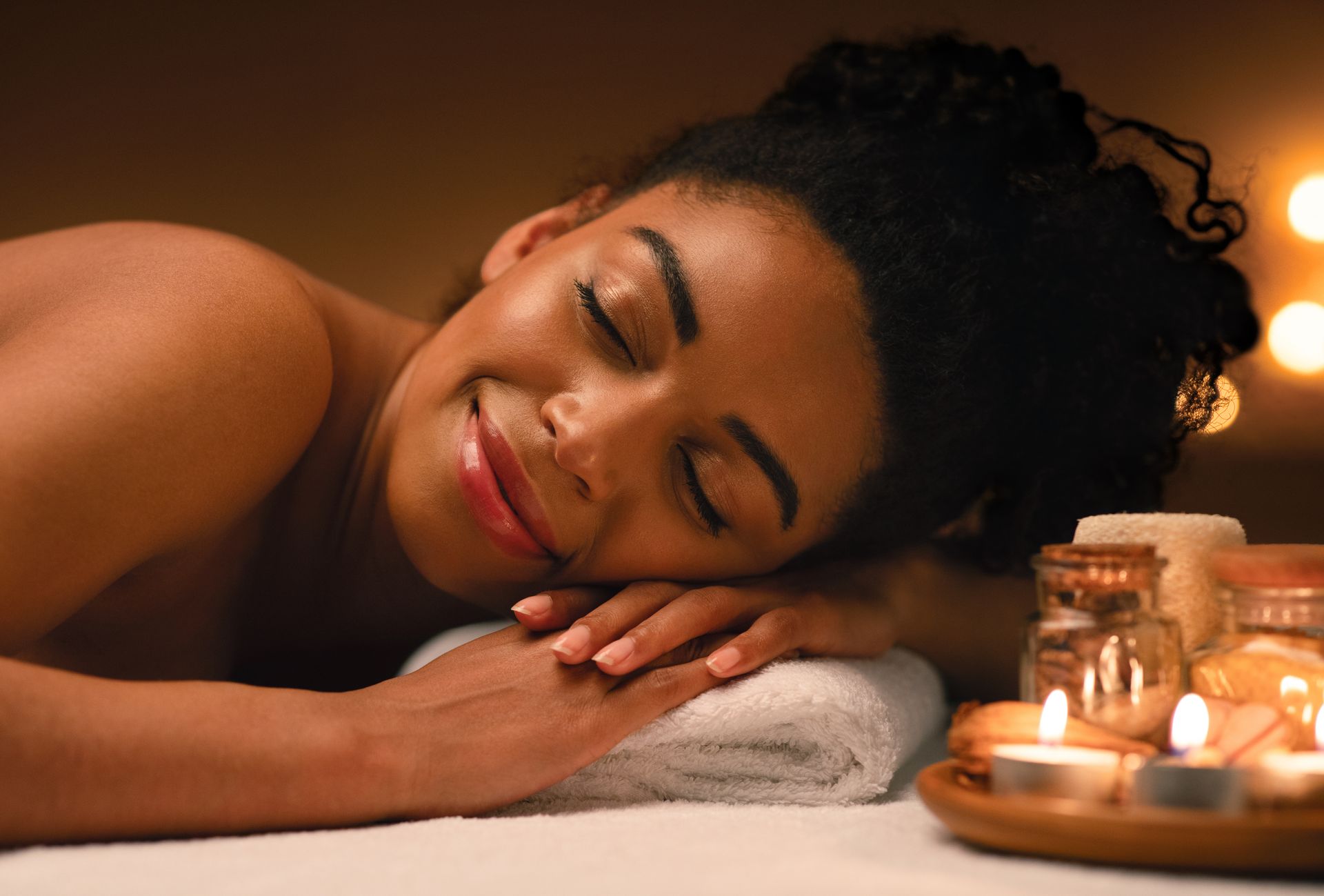 Une femme se repose sur une table de spa, les mains jointes, à côté de bougies allumées et de pots de produits de soin.