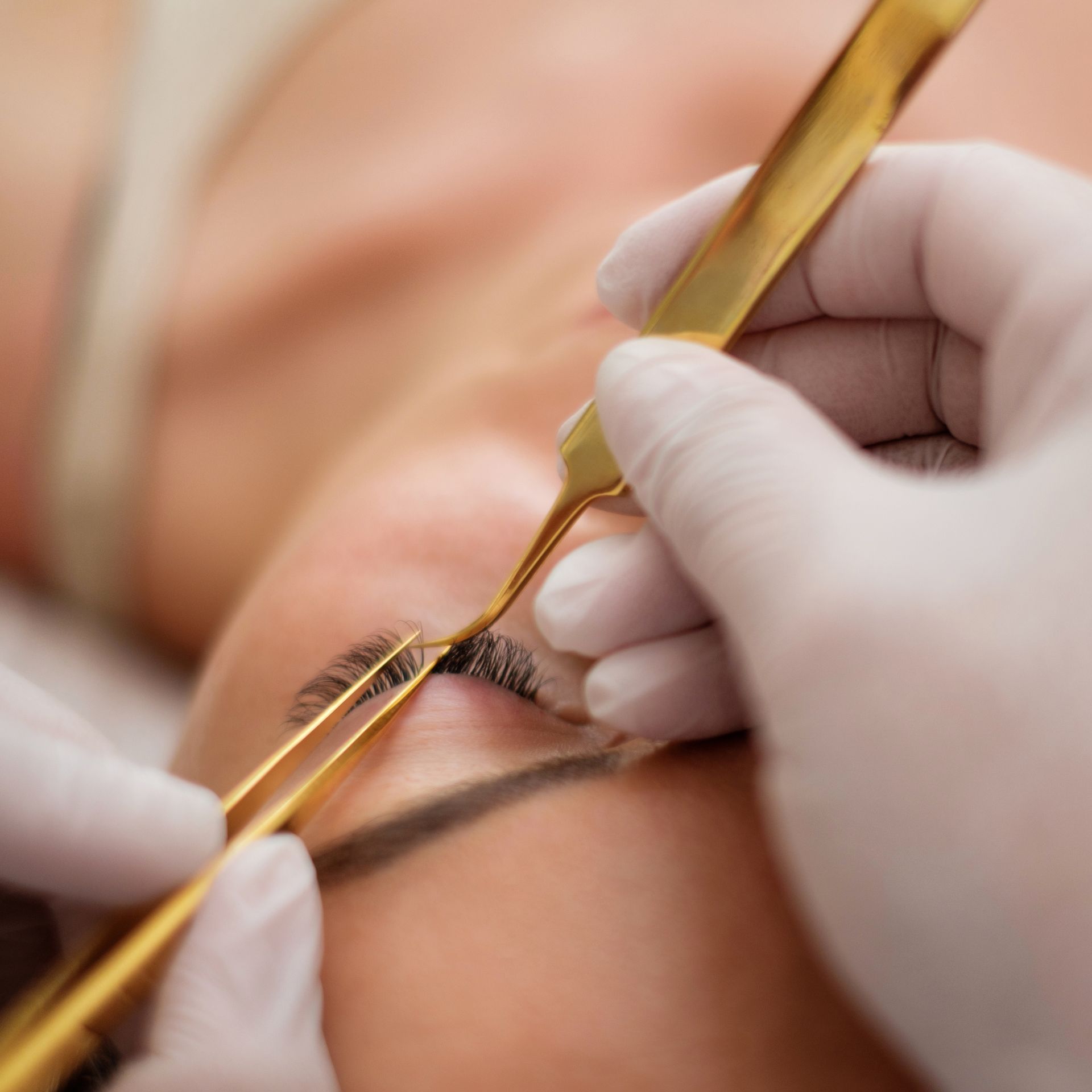 Mains appliquant des pinces à épiler dorées sur une extension de cils posée sur l'œil d'une cliente.