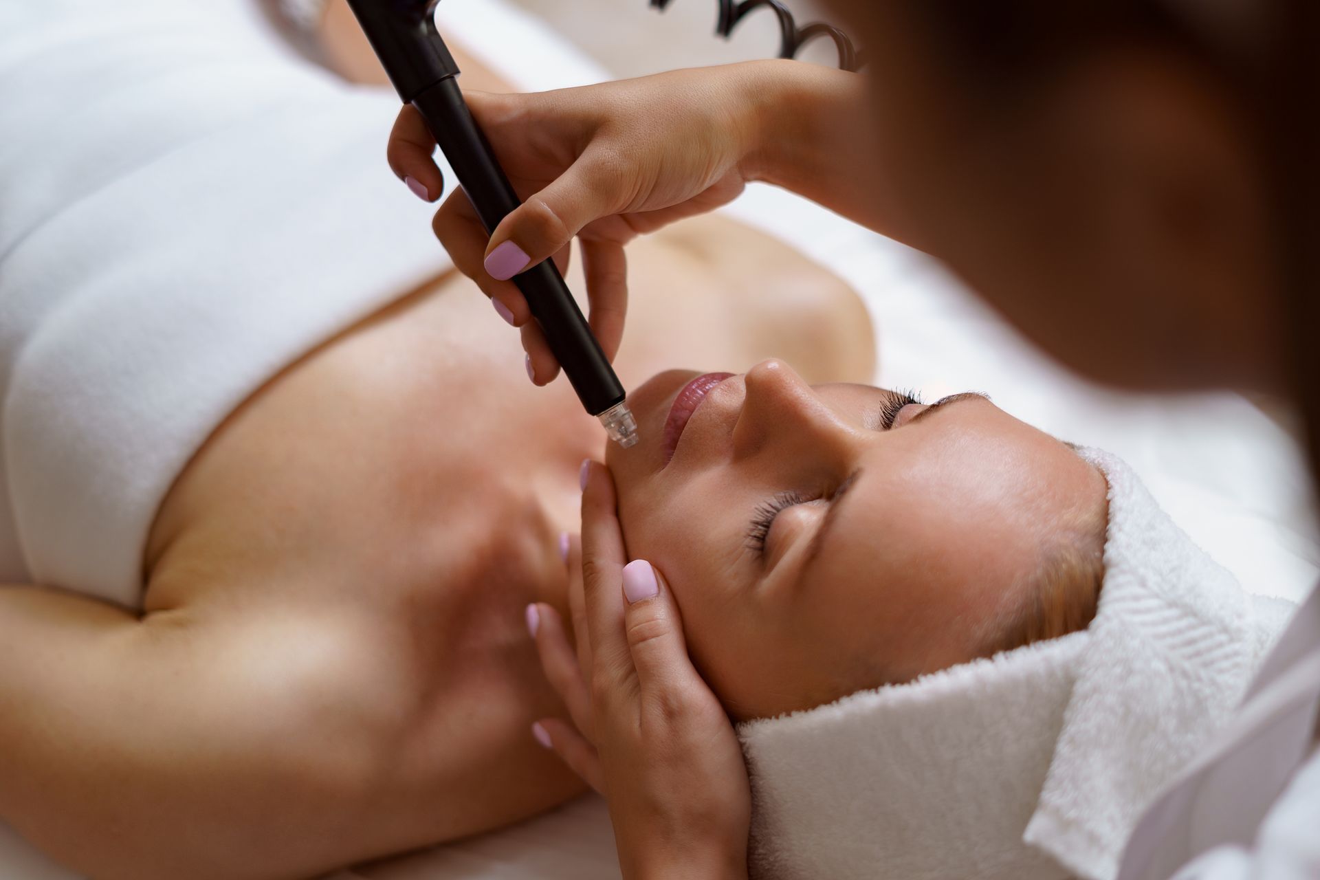 Une esthéticienne applique un soin du visage à une femme allongée, une serviette enroulée autour de la tête.