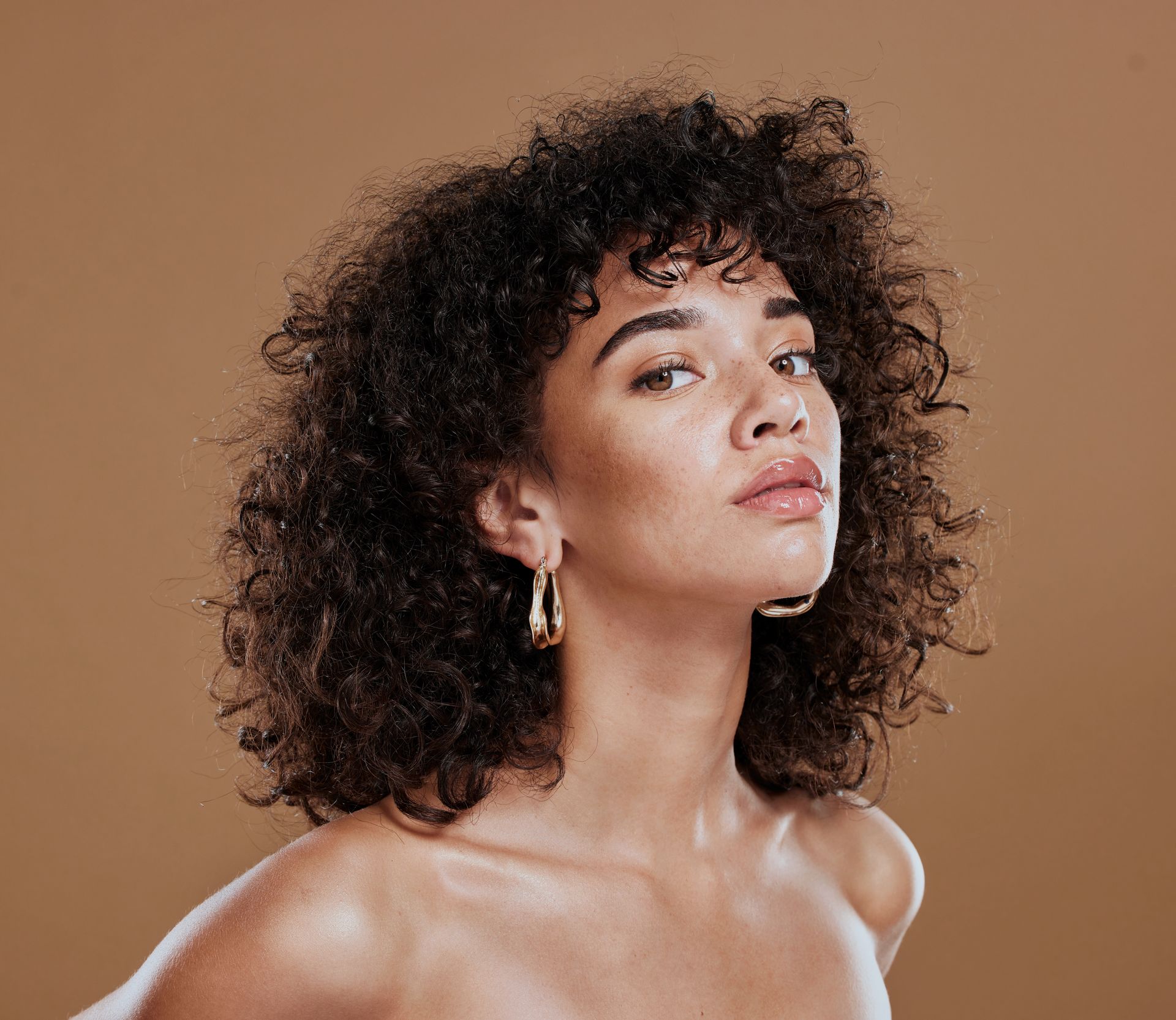 Portrait d'une personne aux cheveux bouclés et aux boucles d'oreilles en or sur fond beige.