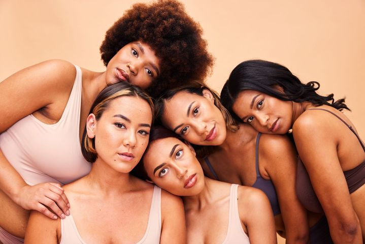 Cinq femmes posent côte à côte sur un fond beige chaud, vêtues de hauts aux tons clairs.