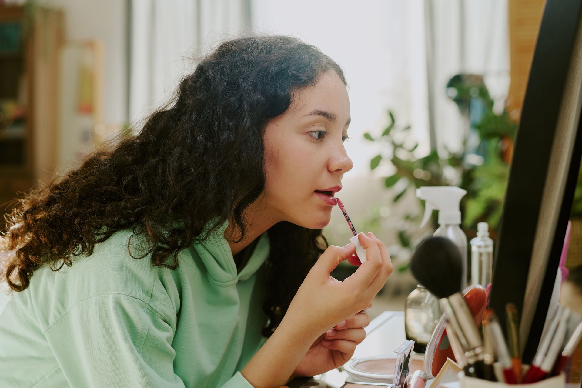 Une femme applique son rouge à lèvres devant un miroir de coiffeuse, avec des pinceaux et des flacons de maquillage.