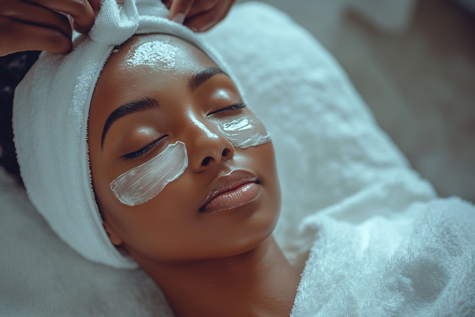 Une personne recevant un soin du visage avec des patchs pour les yeux, allongée sur un lit, enveloppée dans une serviette.