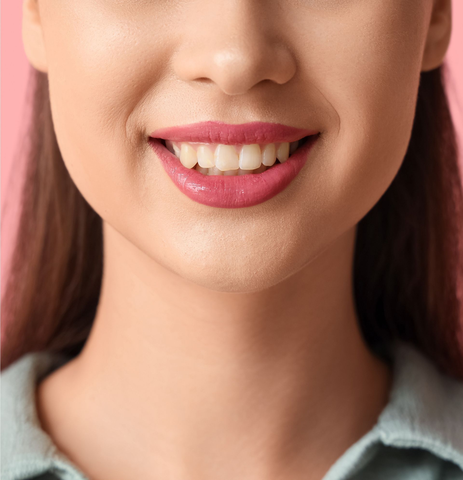 Gros plan sur une bouche souriante avec du rouge à lèvres rose et des dents blanches sur un fond rose pâle.