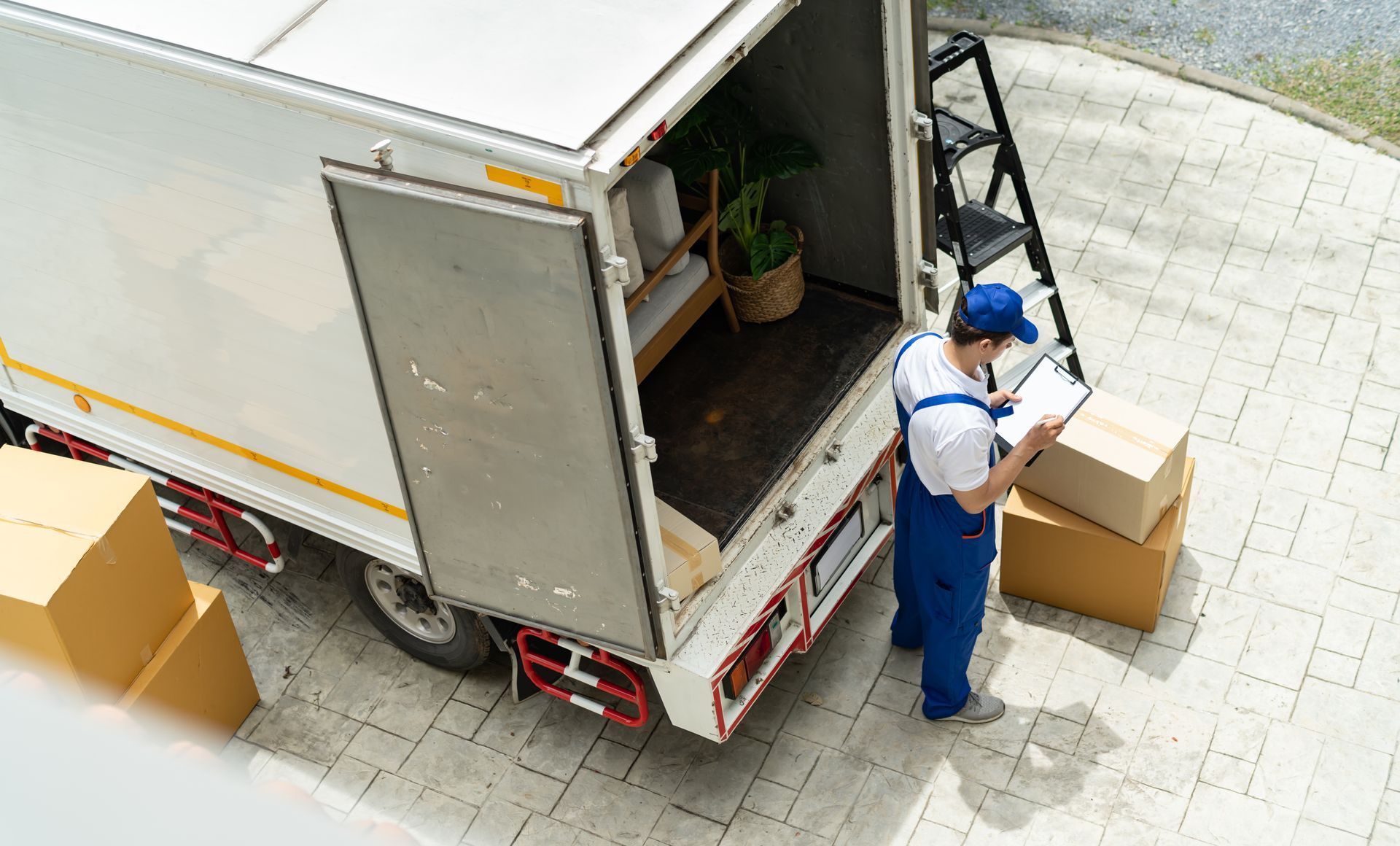 Un livreur en combinaison bleue consulte un bloc-notes près de l'arrière ouvert d'un camion, des cartons empilés autour.