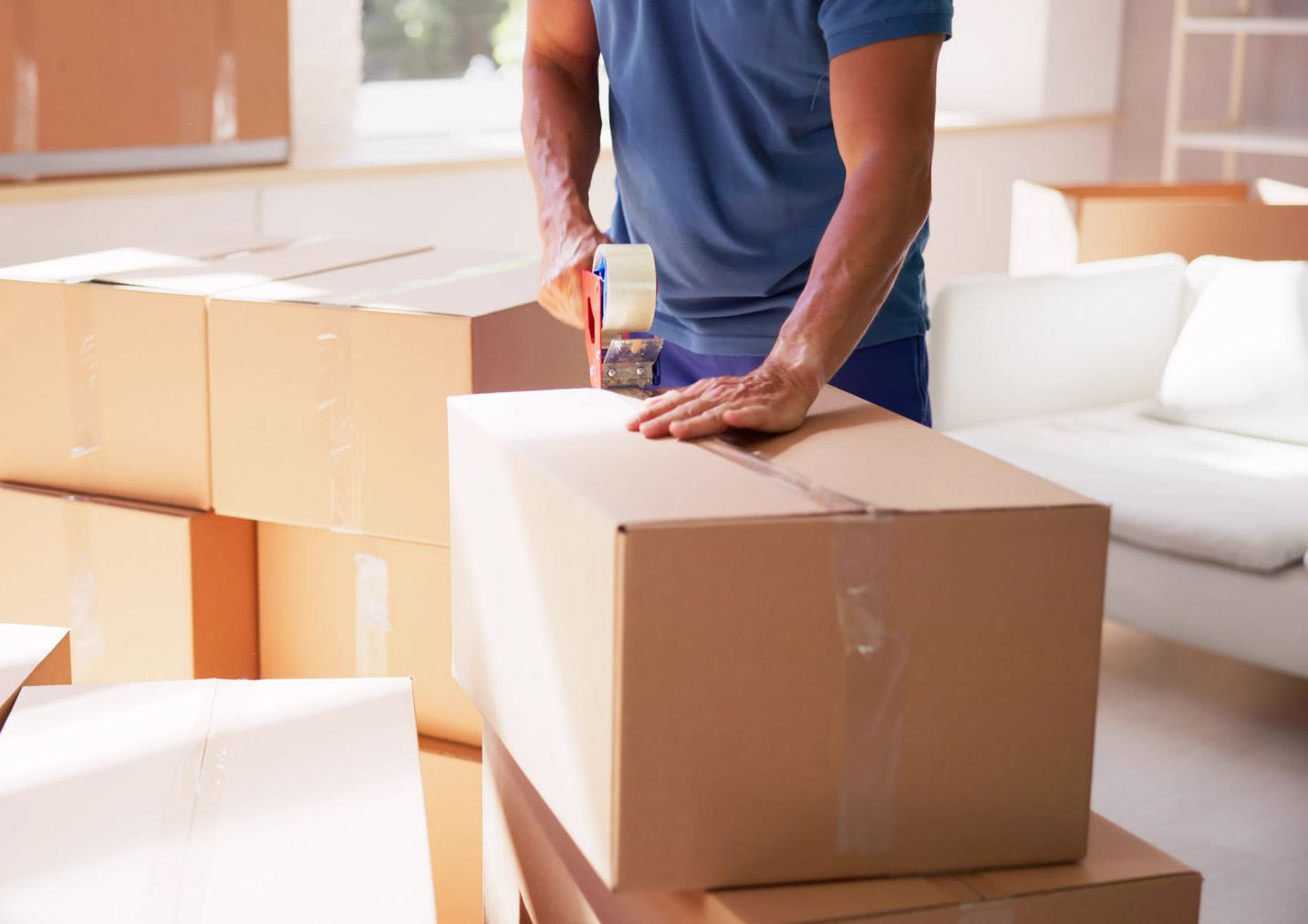 Une personne scotchant un carton, entourée de cartons de déménagement empilés dans une pièce bien éclairée.