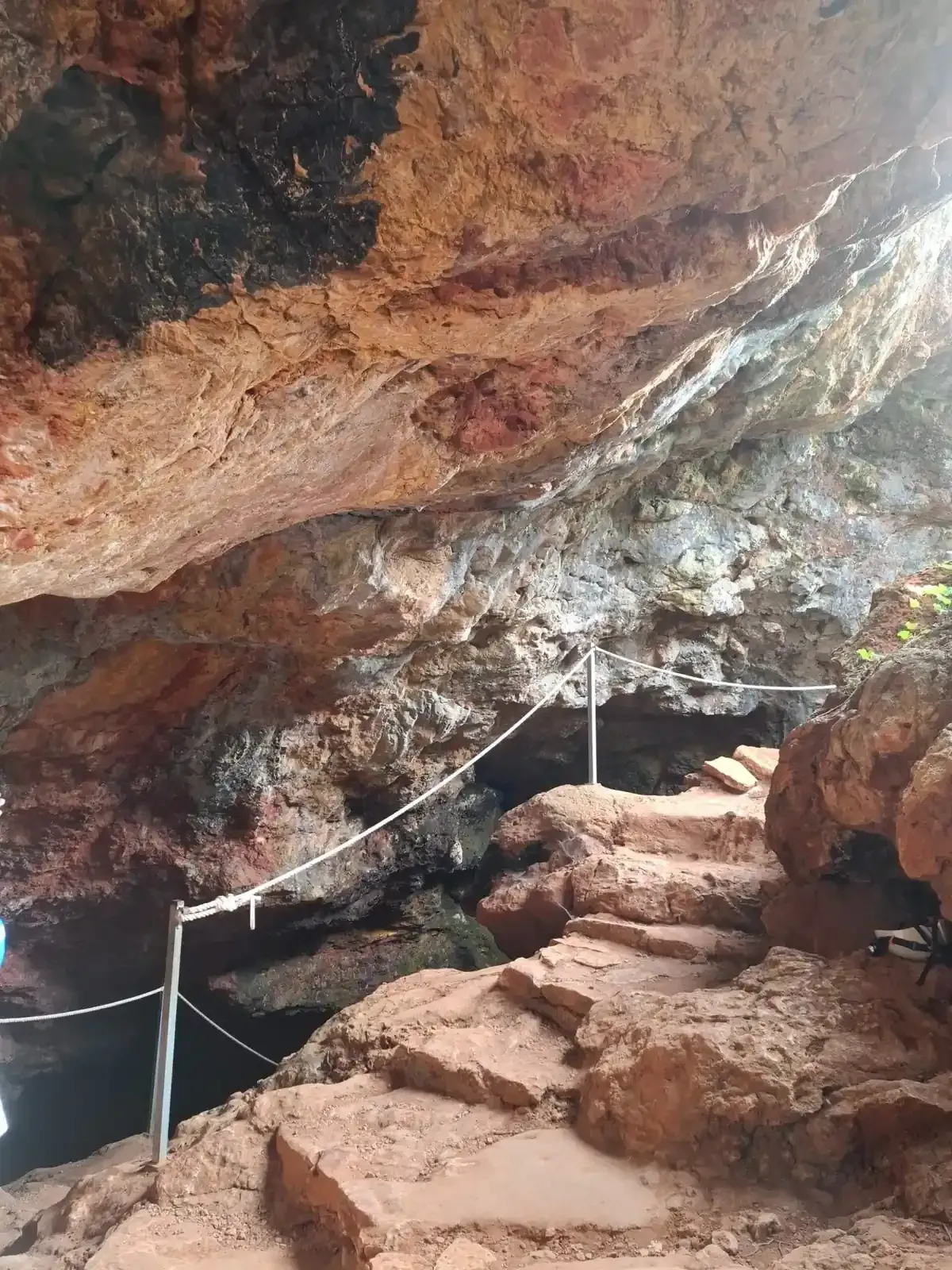 Unas escaleras de piedra conducen a una cueva. Paredes beige y marrones, con pasamanos de cuerda.