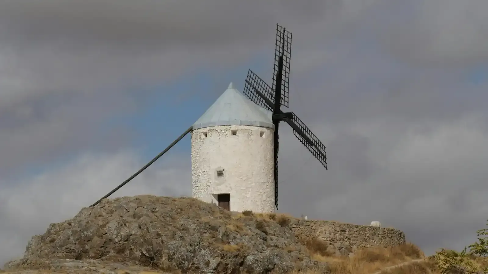 Molino de viento blanco con aspas oscuras en lo alto de una colina rocosa bajo un cielo nublado.
