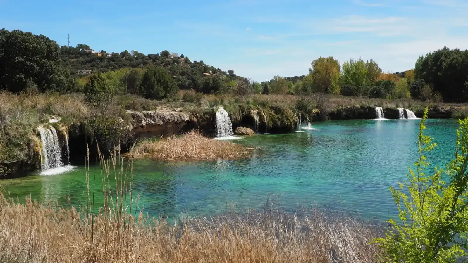 Un lago turquesa con varias cascadas pequeñas bajo un cielo azul. Una exuberante vegetación rodea el agua.