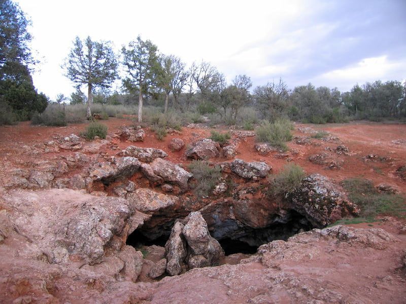 Entrada de la cueva en tierra de color marrón rojizo con rocas dispersas, pequeños árboles y arbustos al fondo bajo un cielo nublado.