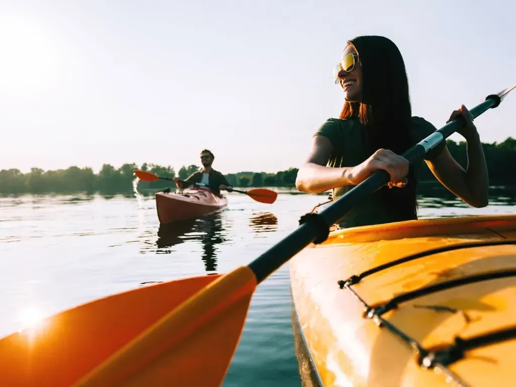 Mujer haciendo kayak con expresión sonriente; otro kayakista al fondo en un lago soleado.