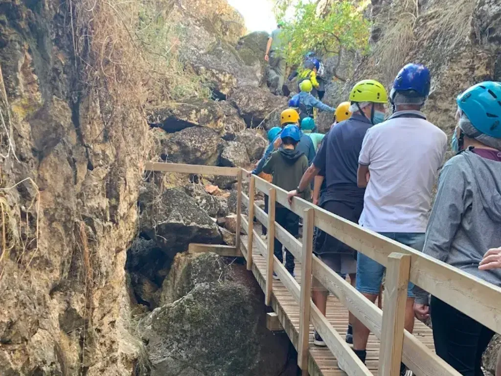 Grupo de personas con cascos caminando sobre un puente de madera en un desfiladero rocoso.