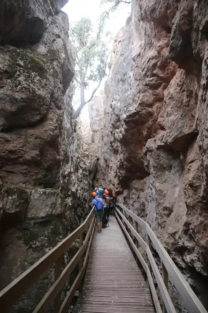 Pasarela de madera a través de un estrecho cañón; grupo de personas caminando; paredes rocosas.