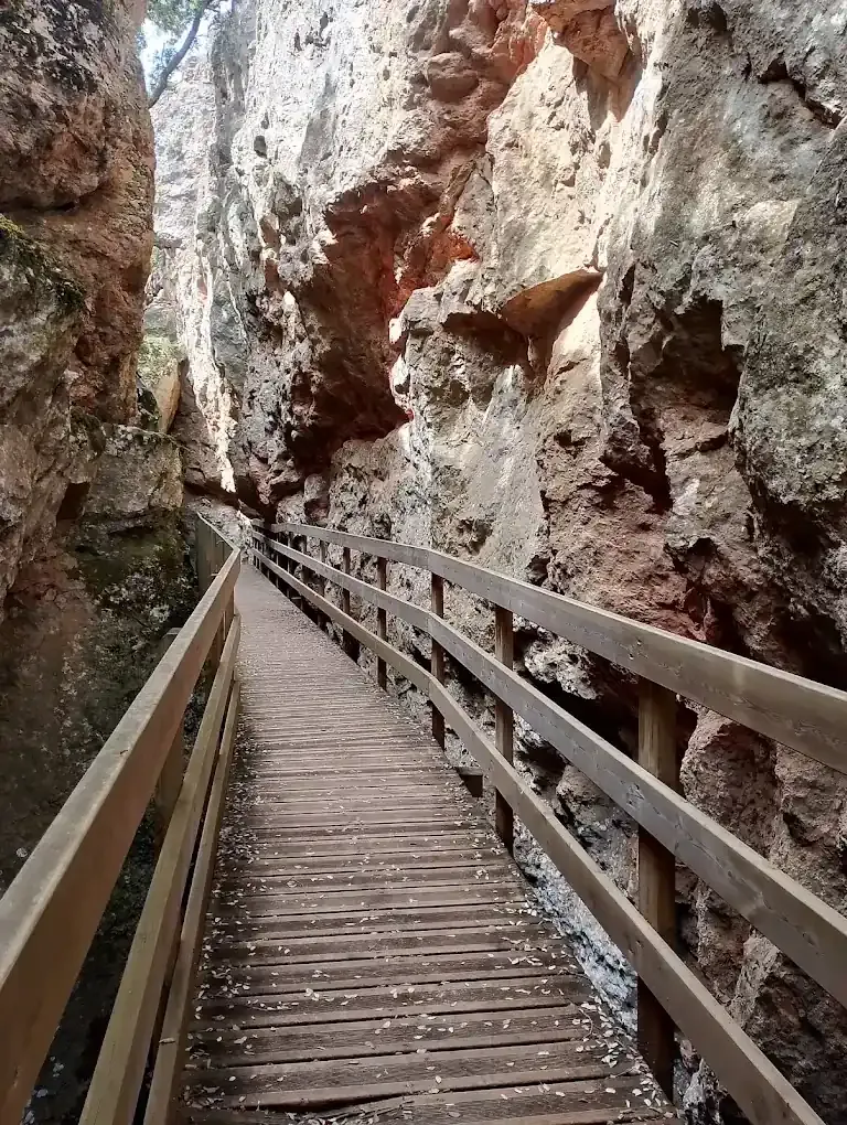 Pasarela de madera que atraviesa un estrecho cañón con paredes de roca rojiza. La luz del sol proyecta sombras.