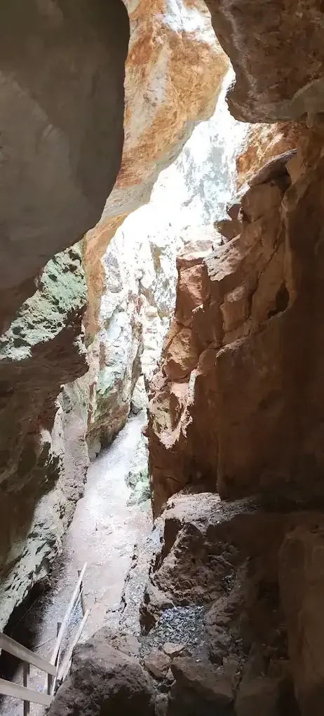 Cueva estrecha con un sendero iluminado por la luz del sol. Se ven paredes rocosas con una barandilla de madera.