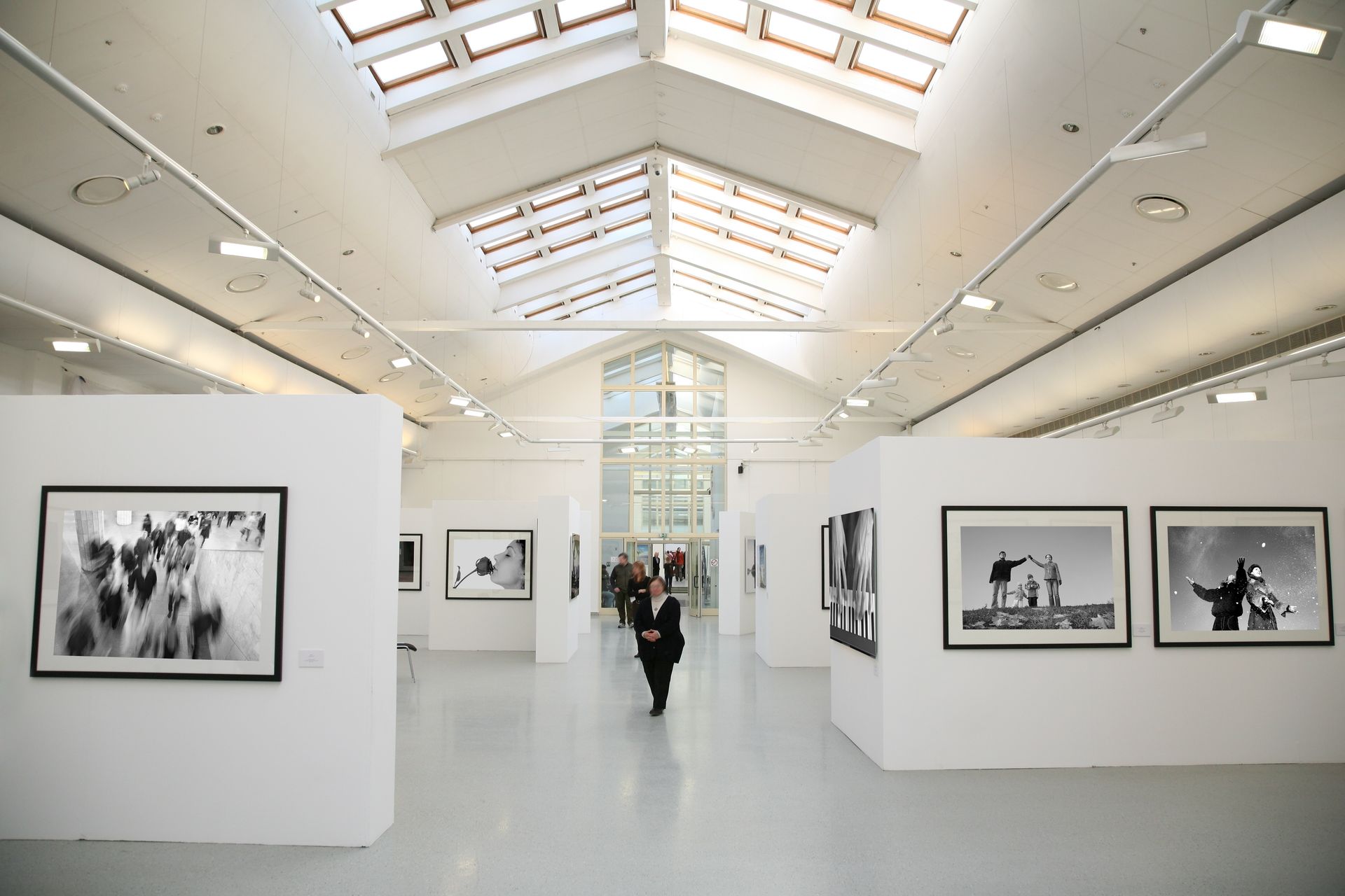 Galerie d'art lumineuse et spacieuse aux murs blancs et aux puits de lumière, présentant des photographies encadrées en noir et blanc. Une personne y déambule.
