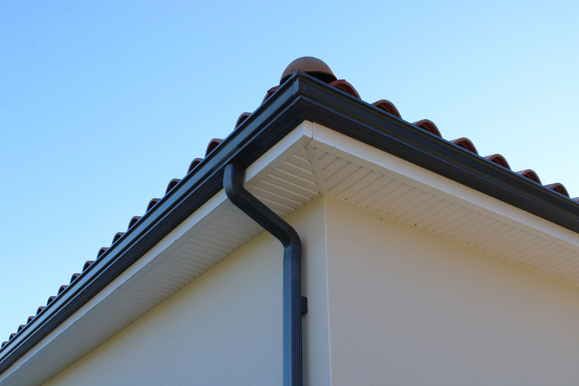 Coin d'un bâtiment avec gouttières sombres, soffite blanc et toit de tuiles rouges sur fond de ciel bleu.