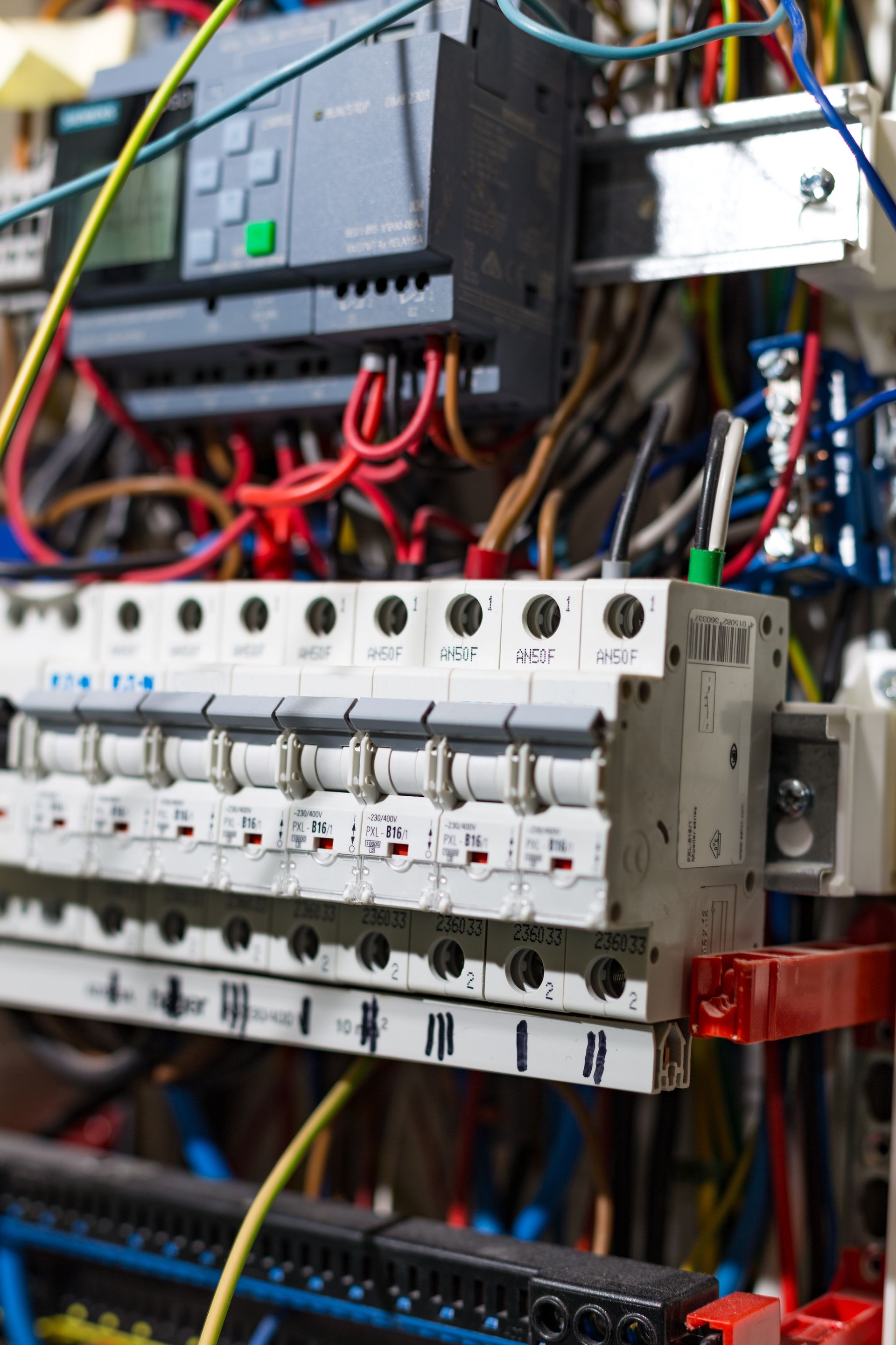 Gros plan sur un panneau de commande électrique avec une unité PLC au-dessus d'une rangée de disjoncteurs et d'un câblage désordonné.