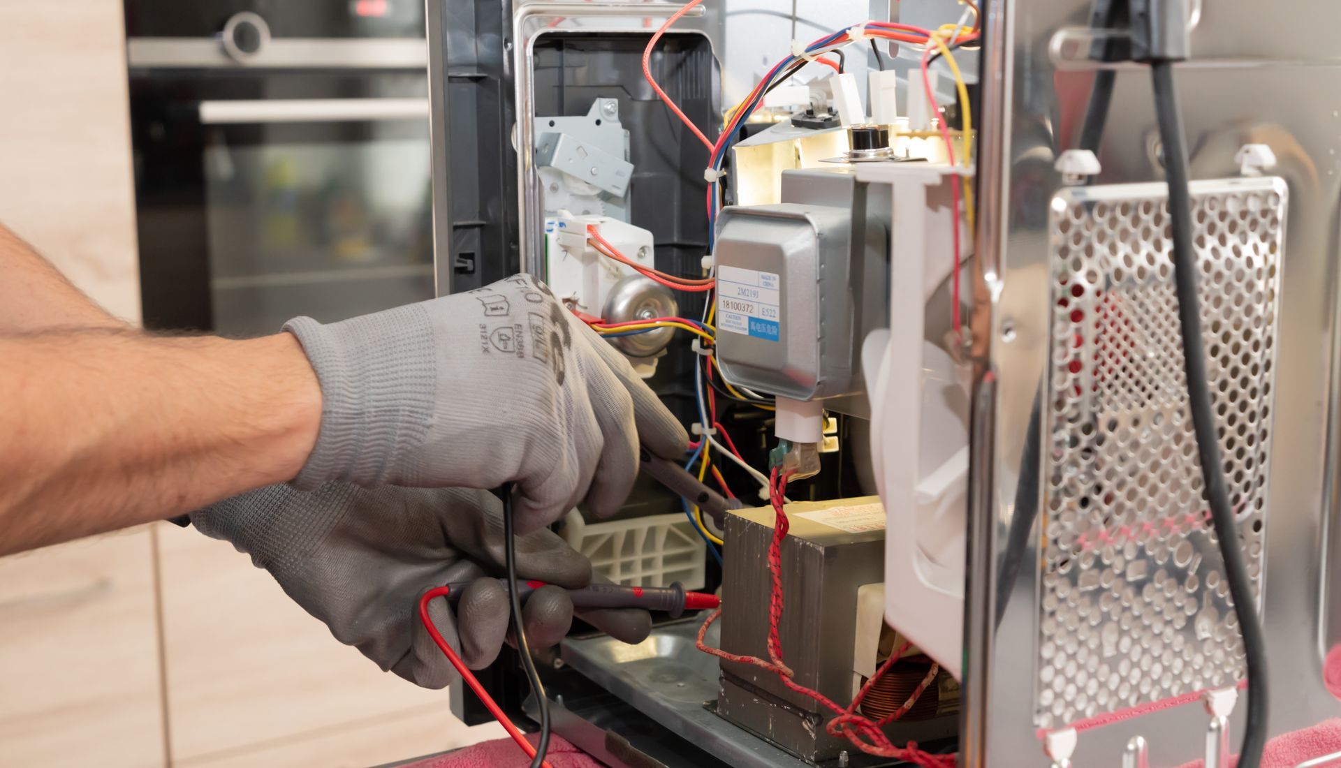 Un technicien portant des gants de travail gris répare les composants électriques internes d'un four à micro-ondes ouvert.