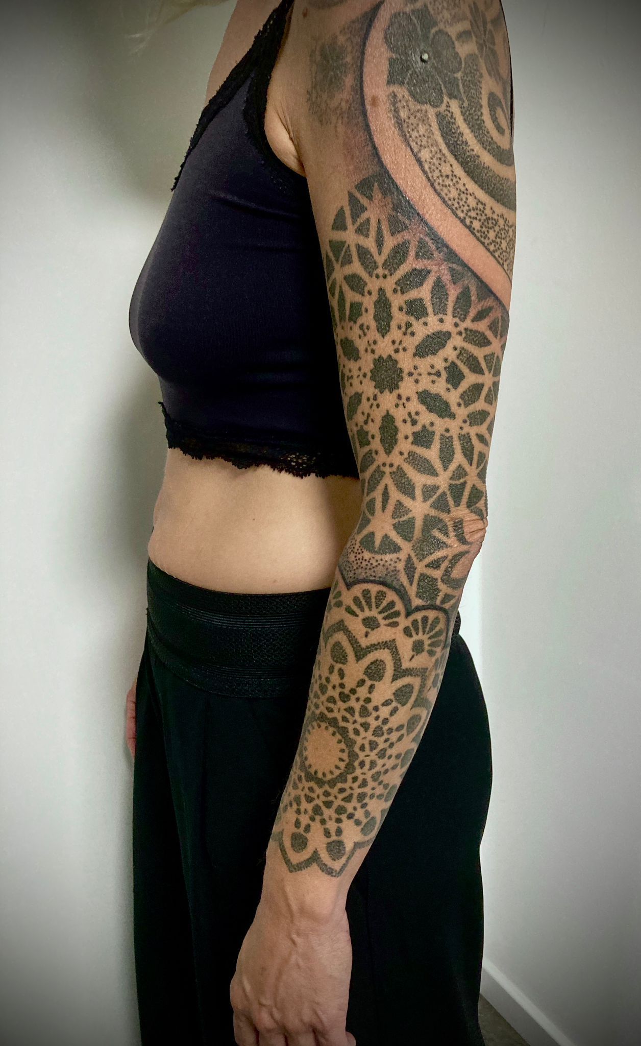 Eine Frau mit einer Tätowierung auf dem Arm trägt ein schwarzes bauchfreies Top und eine schwarze Hose.