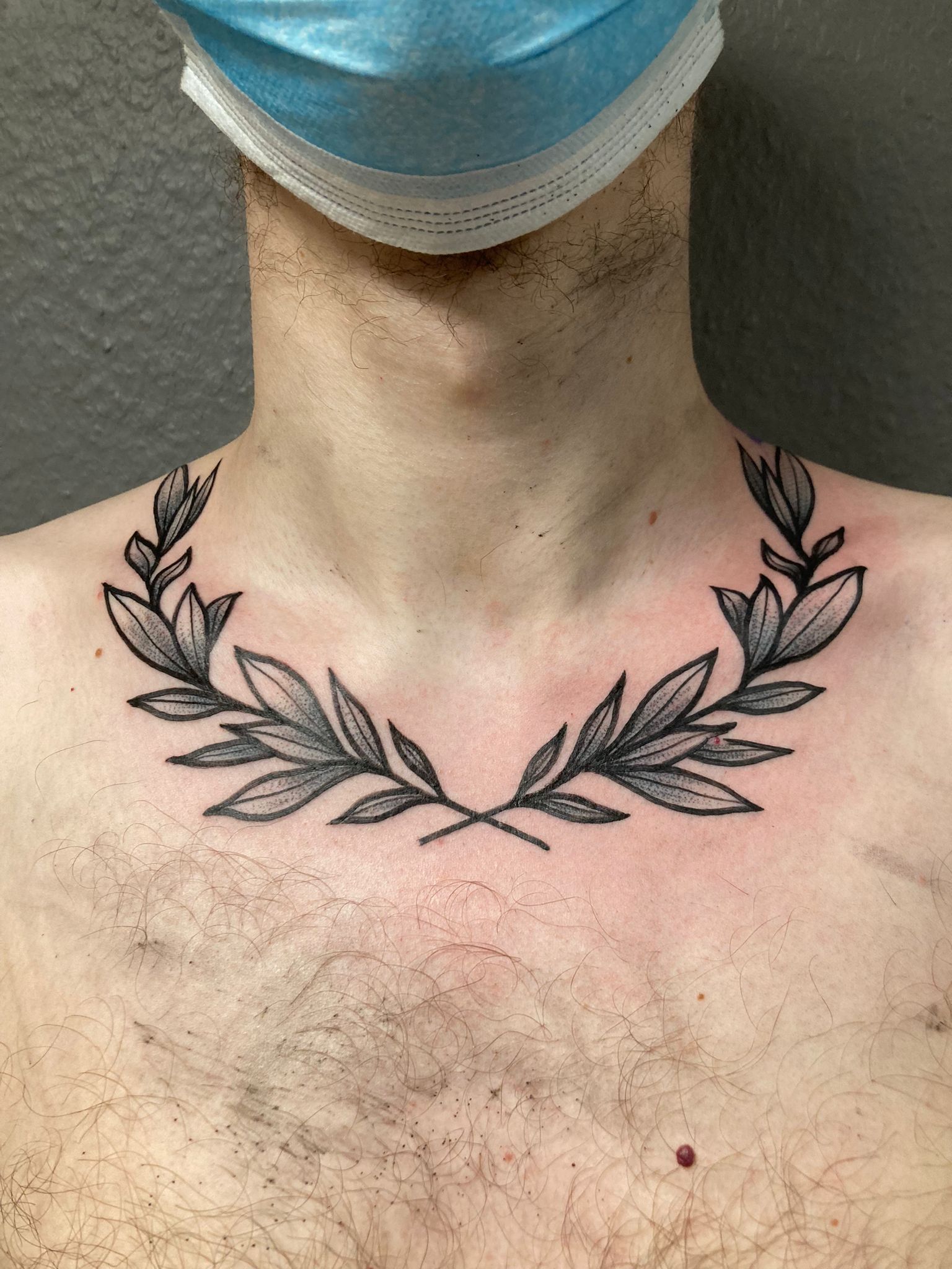 Ein Mann mit einer Maske im Gesicht hat ein Lorbeerkranz-Tattoo auf der Brust.