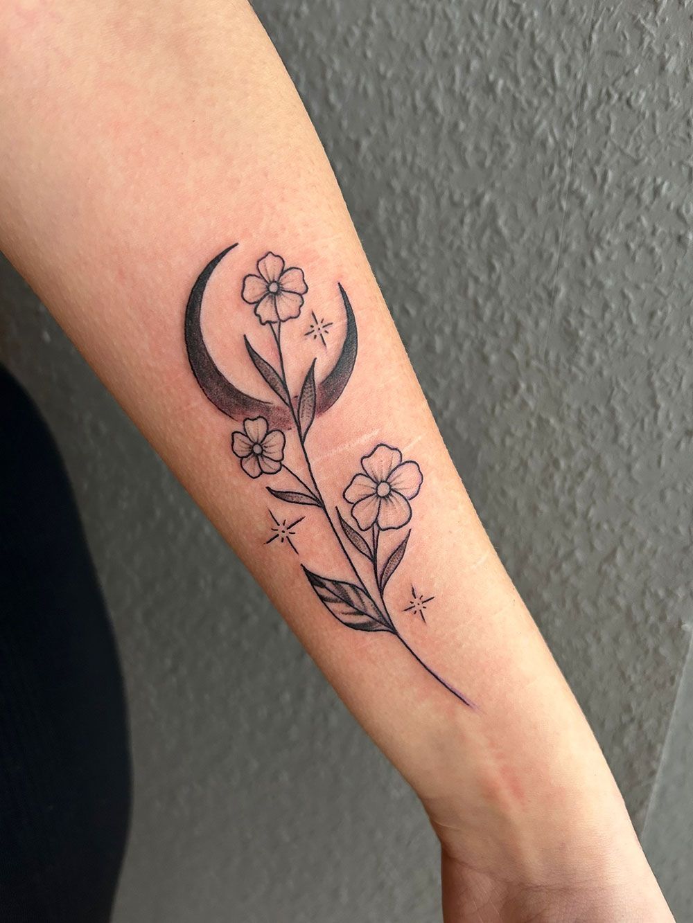 Eine Frau hat eine Tätowierung mit einer Mondsichel und Blumen auf ihrem Unterarm.