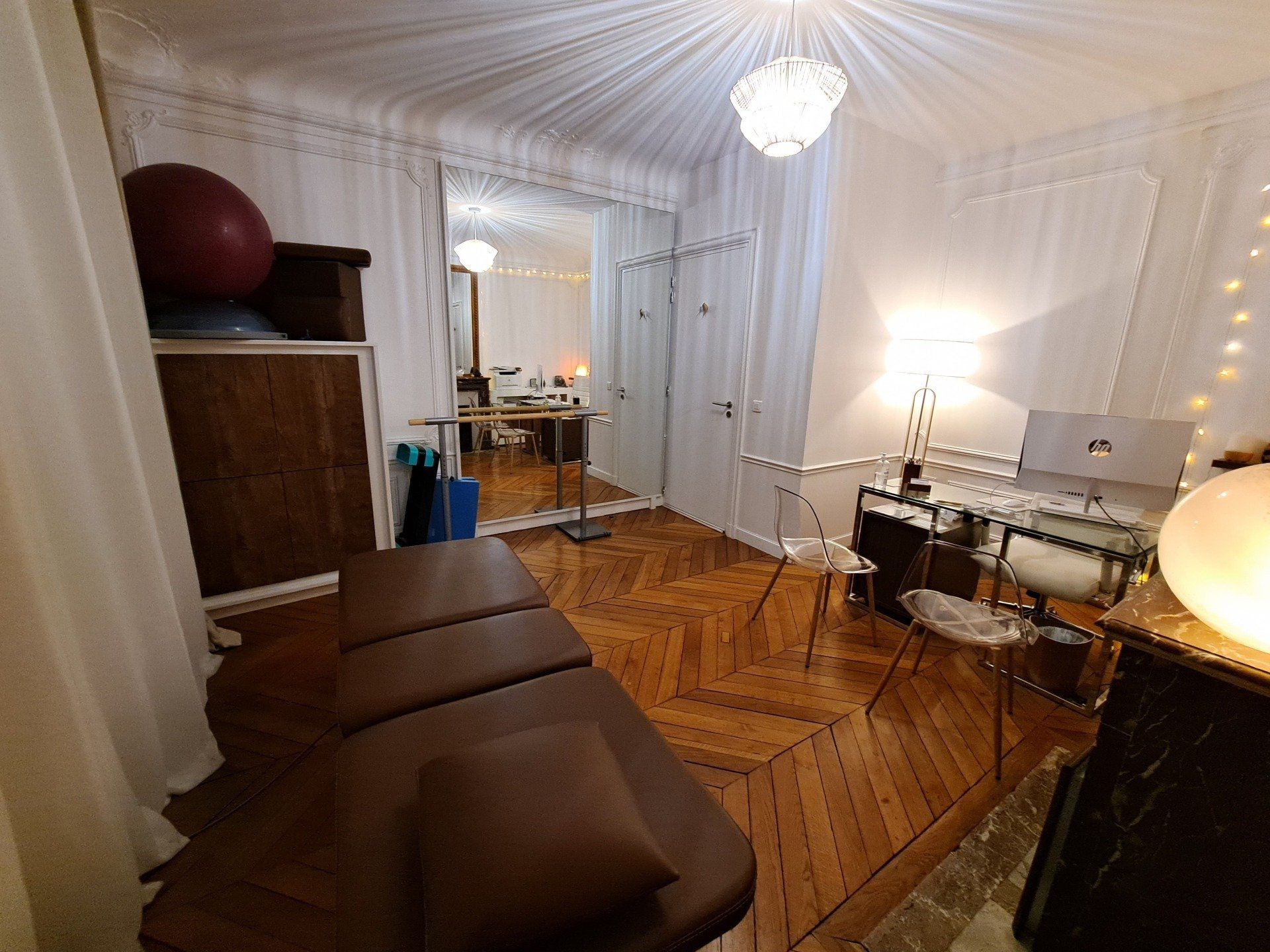 Dans une pièce lumineuse au parquet en chevrons, se trouve une table de massage, un bureau, un grand miroir et un ballon d'exercice rouge.