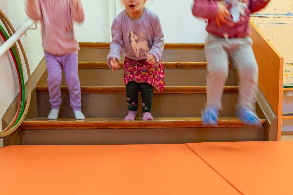 Drei Kinder springen eine Treppe hinunter auf eine orangefarbene Matte.
