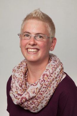 Frau mit kurzen blonden Haaren, Brille und gemustertem Schal, lächelt.