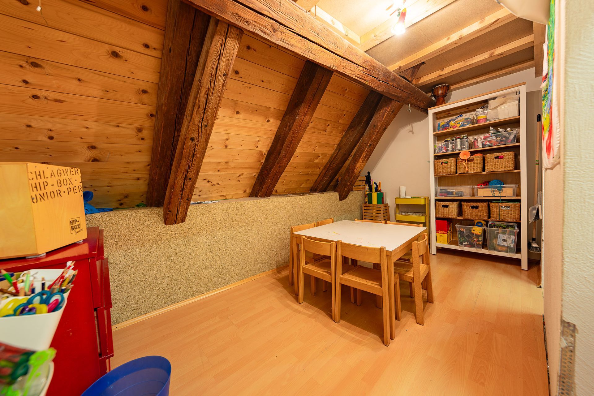 Gemütliches Dachgeschoss-Spielzimmer mit Holzbalken, kleinem Tisch und Stühlen sowie einem mit Spielzeug gefüllten Bücherregal.