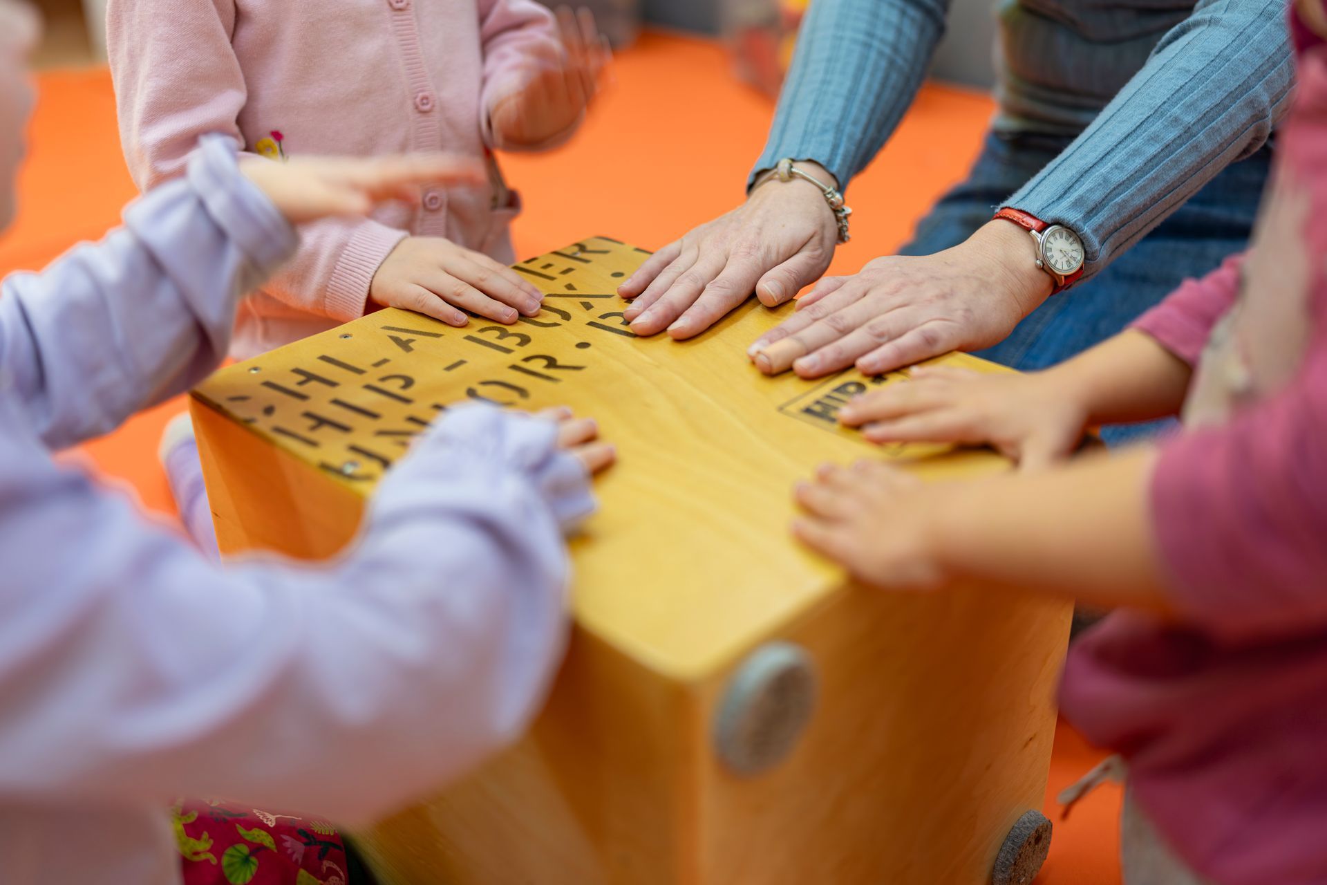 Drei kleine Kinder und ein Erwachsener berühren einen Karton auf einer orangefarbenen Matte.