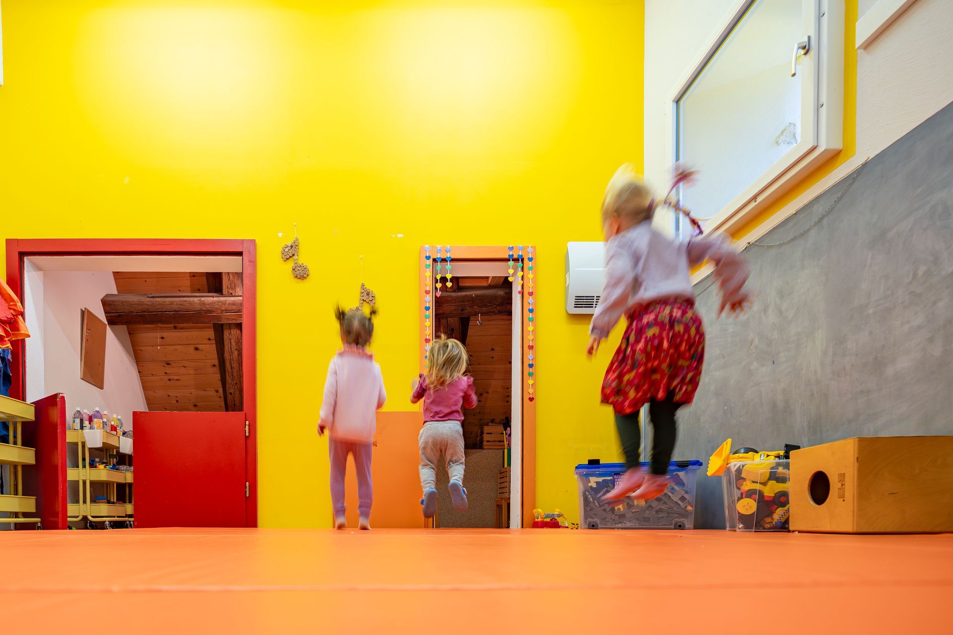 Eine weisse Babyhängematte hängt in einem Kinderzimmer an einem Holzbalken; ein Kinderbett und andere Gegenstände sind zu sehen.
