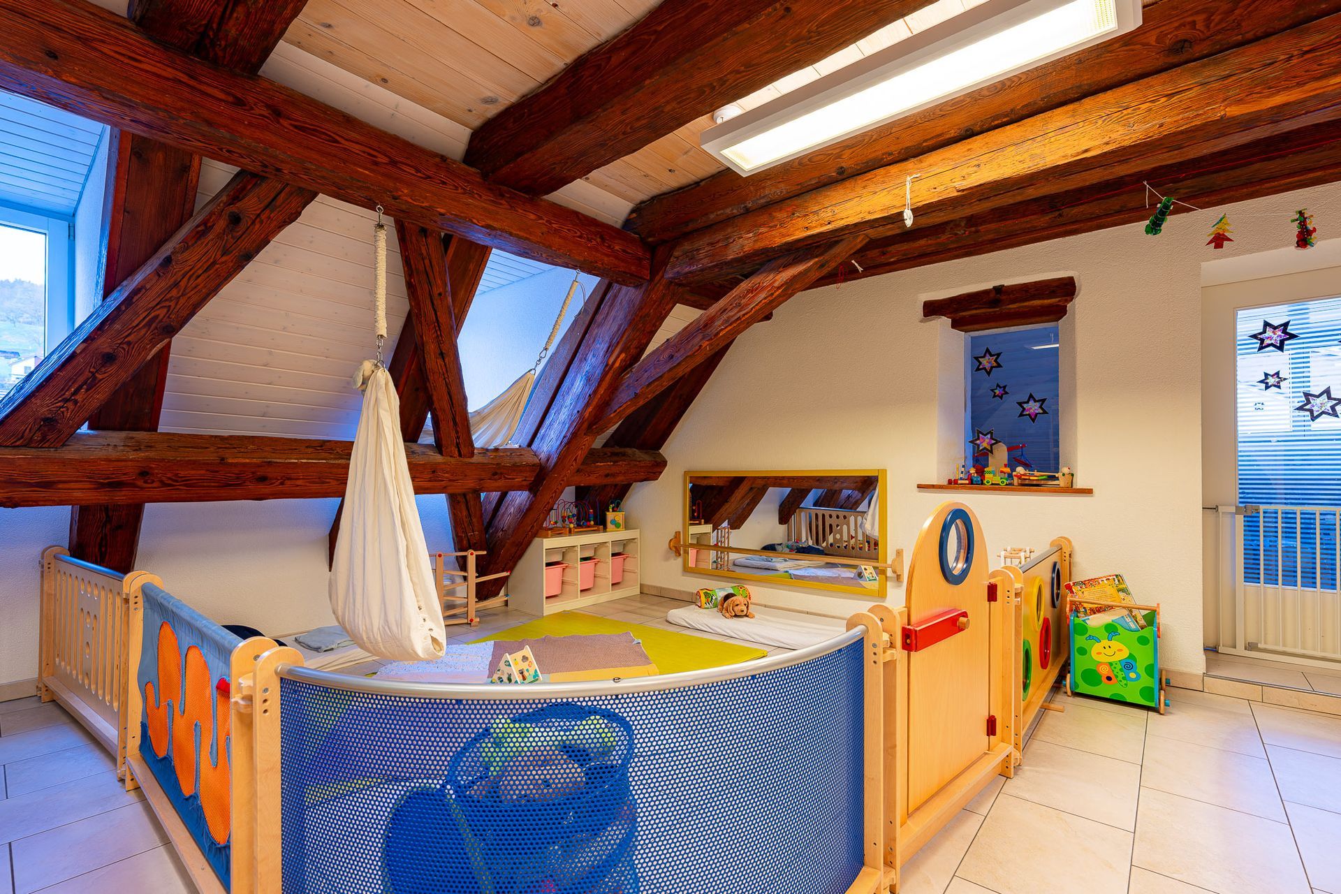 Spielzimmer mit Holzbalken, blau-orangem Laufgitter, Schaukel und kleinem Fenster mit Sternen.