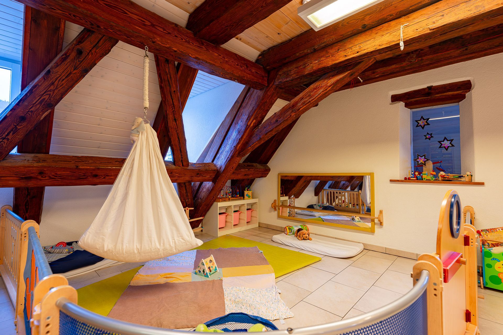 Spielzimmer für Kinder mit Holzbalken, einer Hängeschaukel, Kinderbetten, einem bunten Teppich und Spielzeug.