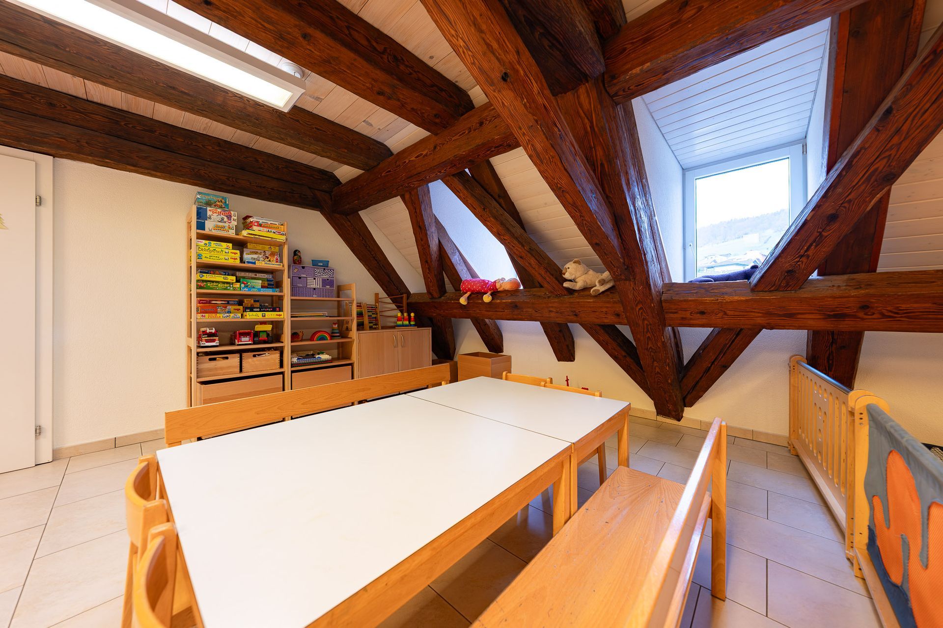 Klassenzimmer mit Holzbalken, weißem Tisch, Holzbänken, Regalen mit Spielzeug und einem Fenster.