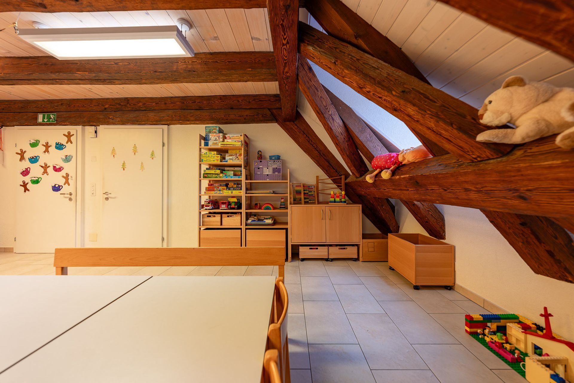 Spielzimmer mit Holzbalken, Regalen, Tisch, Spielzeug und einem Stoffbären.