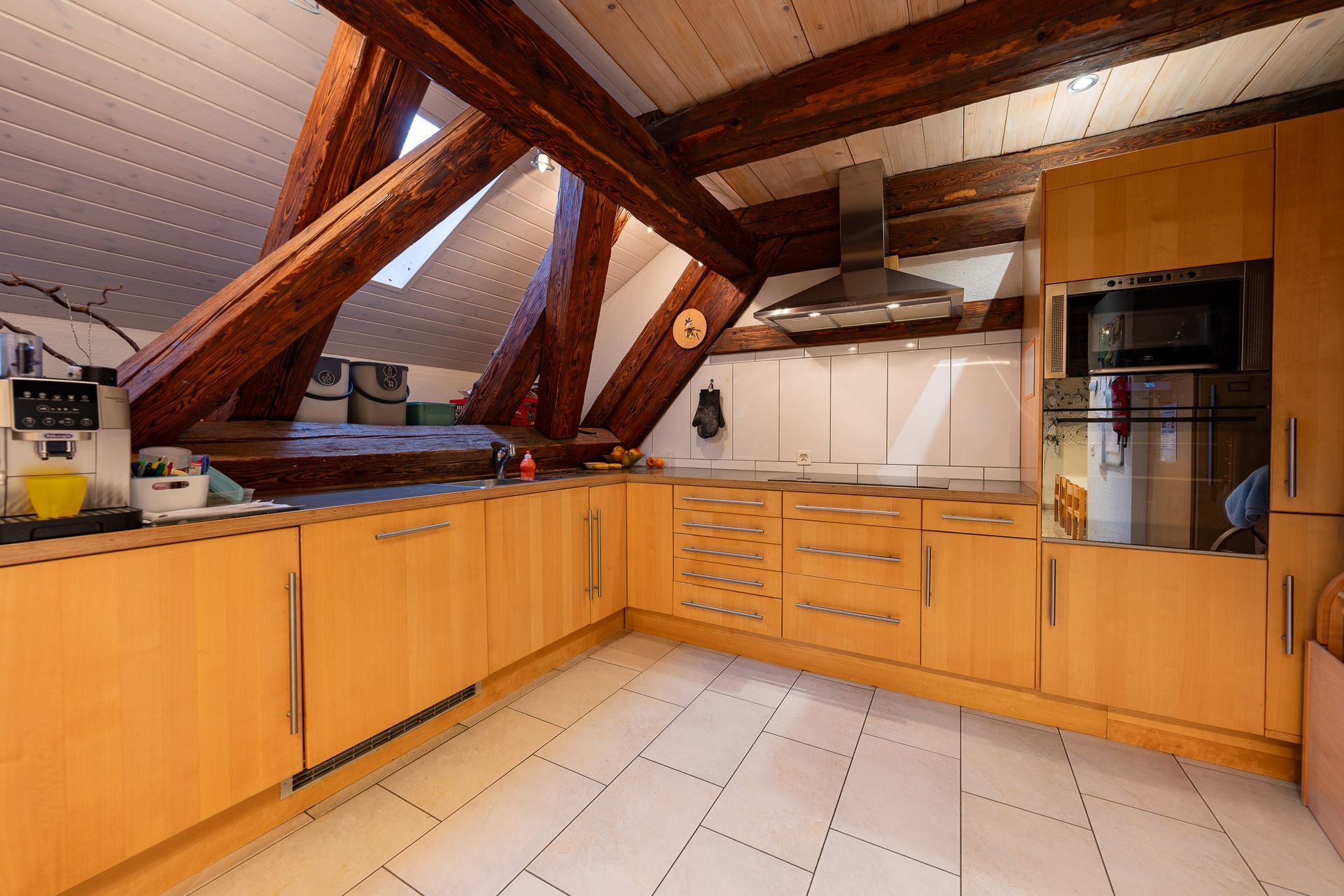 Küche mit Holzbalken, hellen Holzschränken, Einbaugeräten und weißem Fliesenboden.