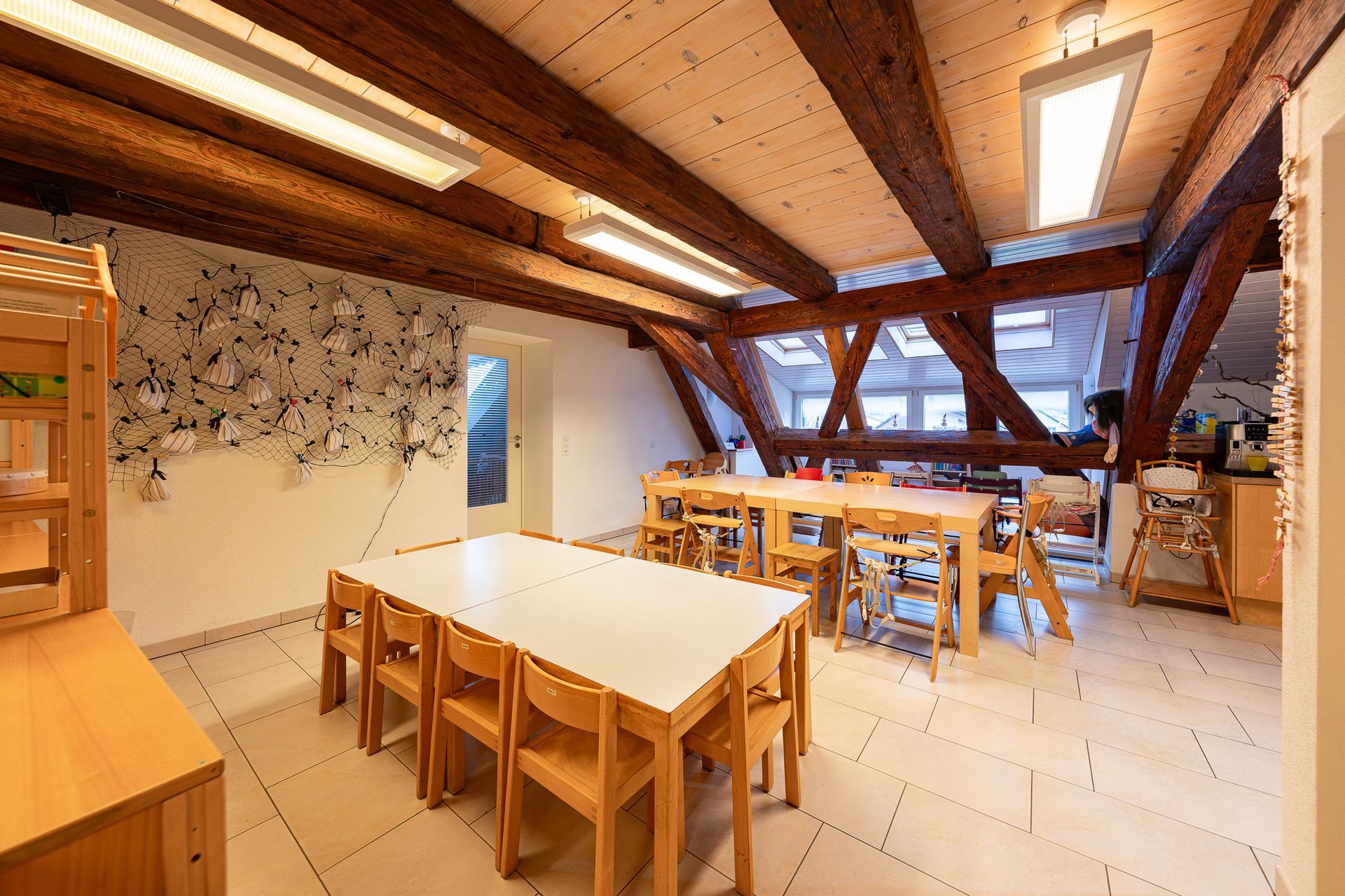 Ein helles, luftiges Klassenzimmer mit Holzbalken und -tischen. Es enthält mehrere Stühle, Fenster und Regale.