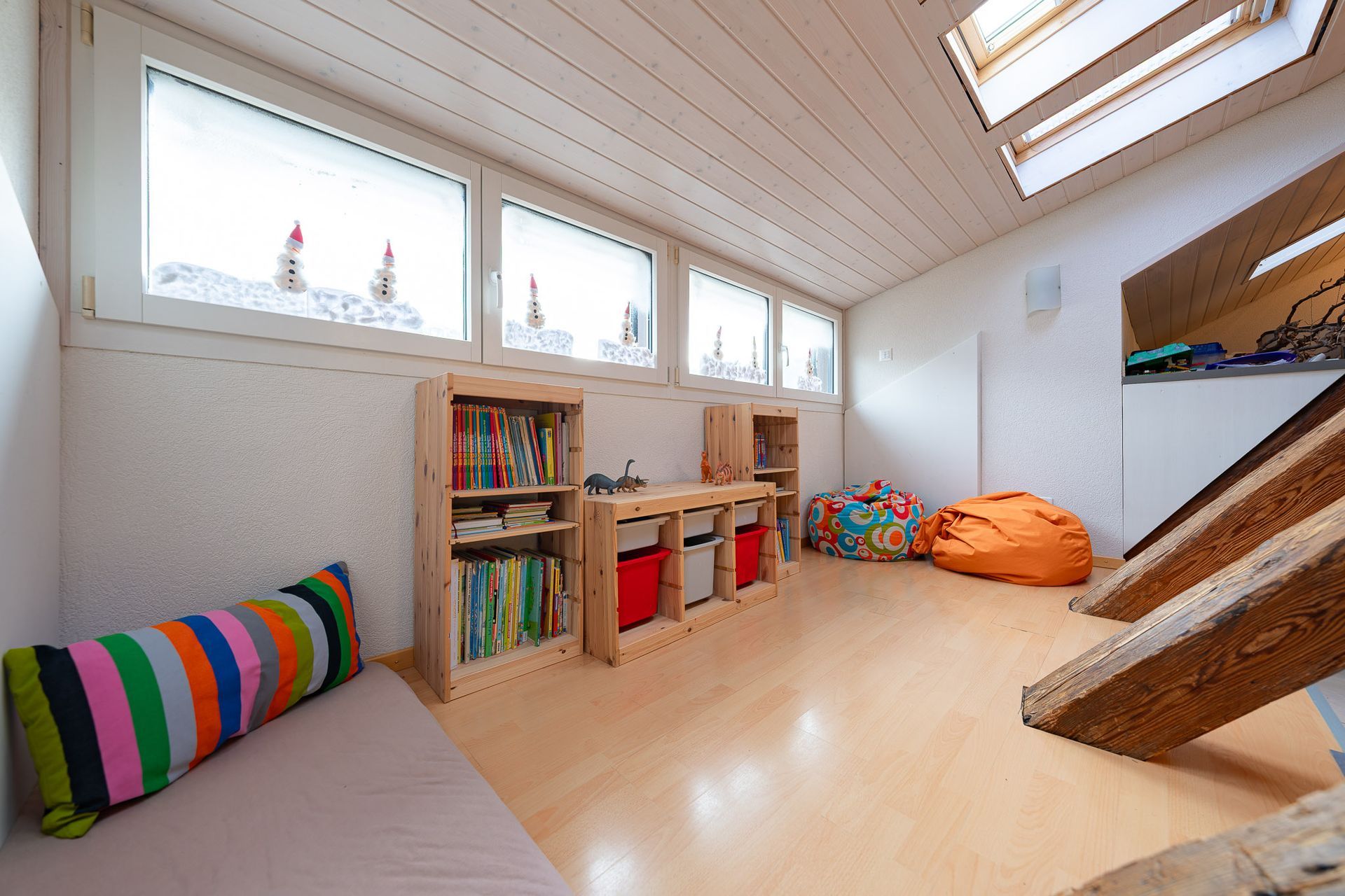 Spielzimmer mit Holzmöbeln, Büchern und bunten Sitzsäcken; schräge Decke mit Fenstern.