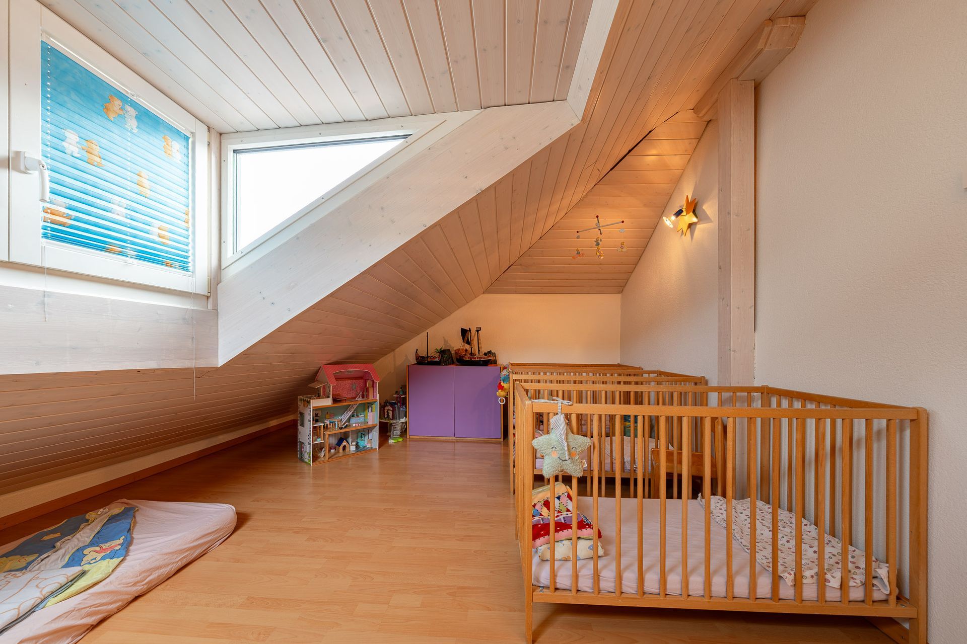 In einem hell erleuchteten Raum mit schräger Decke, möglicherweise einer Kindertagesstätte oder einem Kinderzimmer, stehen hölzerne Kinderbetten.