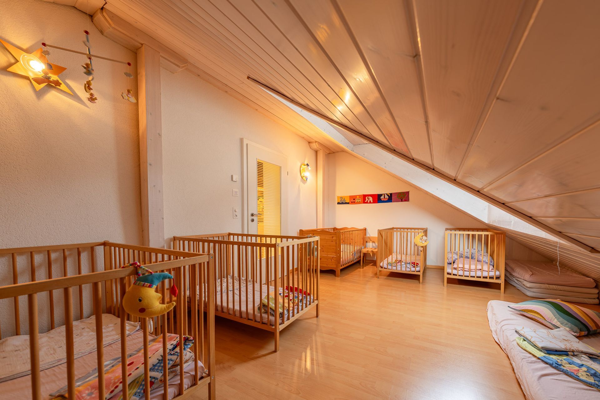 Kinderzimmer mit Holzkinderbetten unter schräger Decke. Sanftes Licht, Holzboden und eine Bodenmatte sind zu sehen.