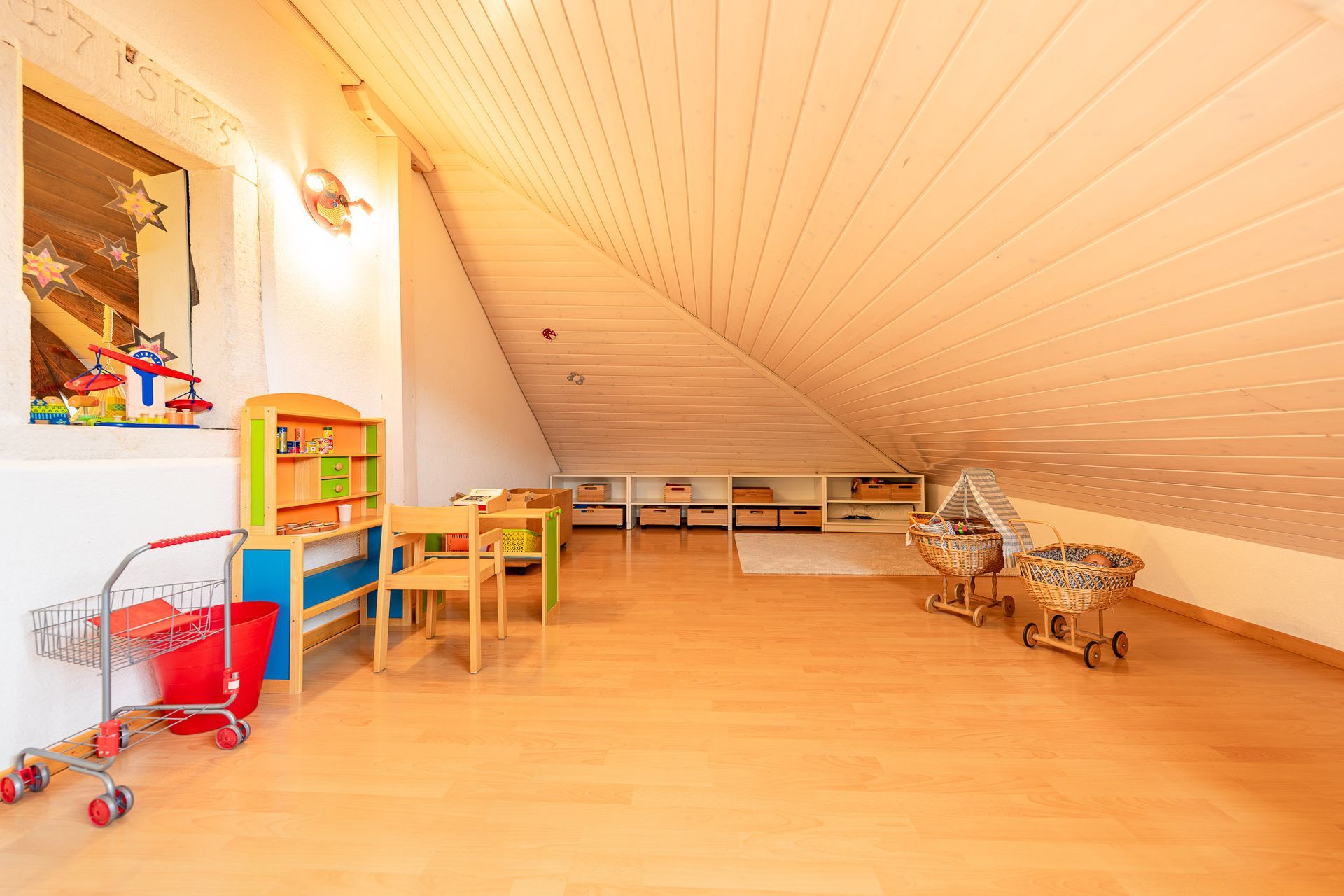 Hell erleuchtetes Dachgeschoss-Spielzimmer mit Holzboden, Dachschräge und Kinderspielzeug.