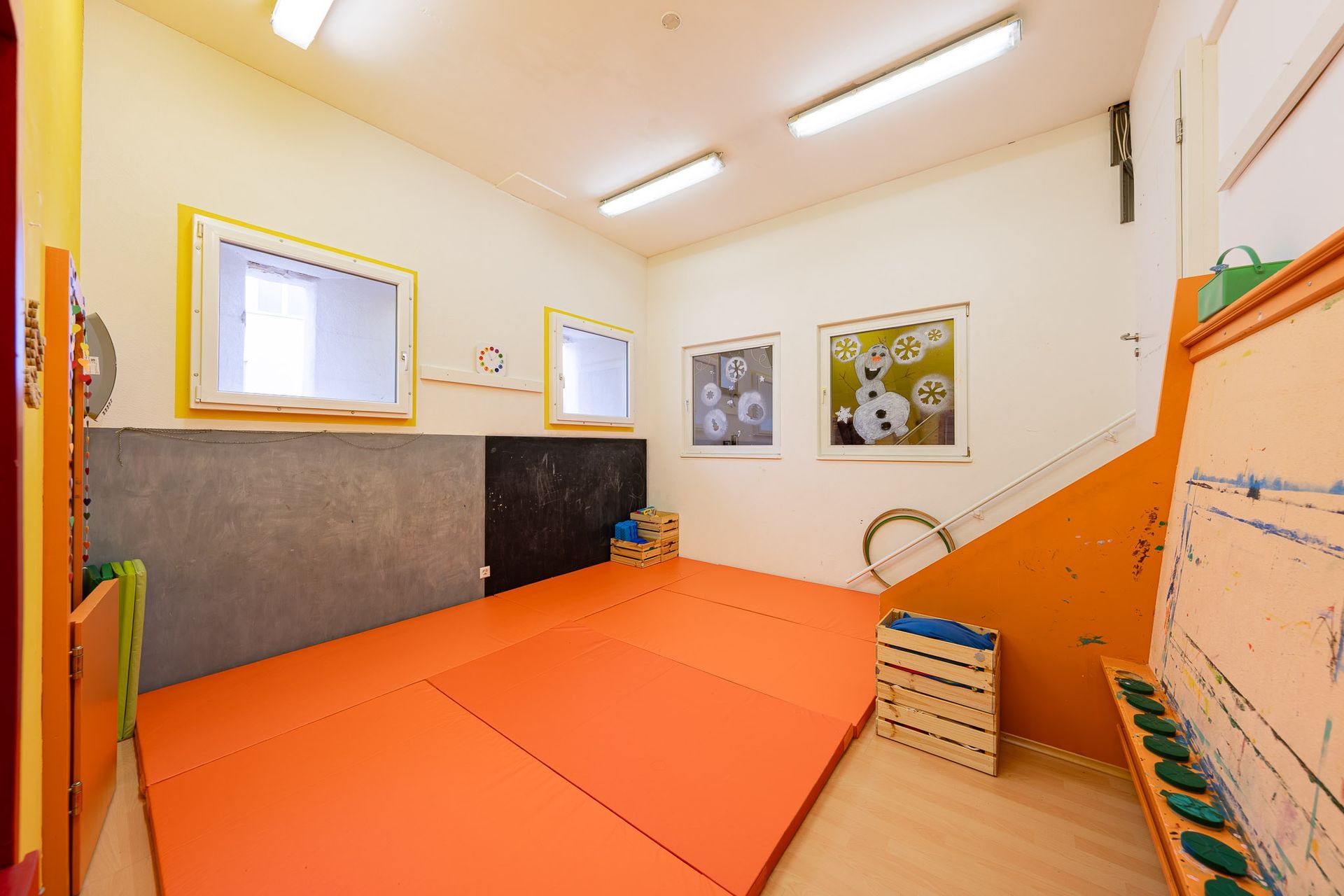 Kinderspielzimmer mit orangefarbenen Matten, Wandbildern und einer Kletterwand.