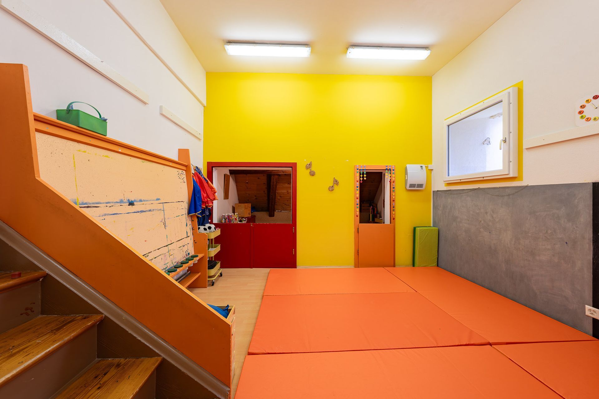 Turnraum mit orangefarbenen Matten, Treppe und gelb-weißen Wänden. Zwei Türen und ein Fenster sind zu sehen.
